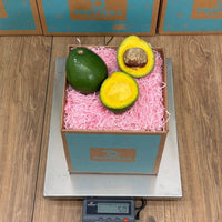 Thumbnail for Tropical Fruit Box 2 ripe avocados, one whole and one halved, in a box with pink crinkle paper on a scale.