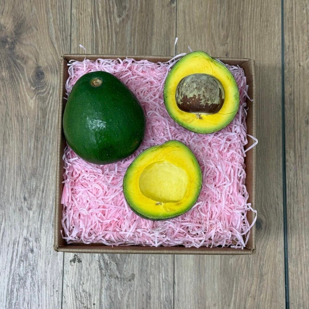 Tropical Fruit Box 2 ct BOGO avocados, one whole and two halves showing bright yellow flesh and large brown pits, nestled in pink crinkle paper.