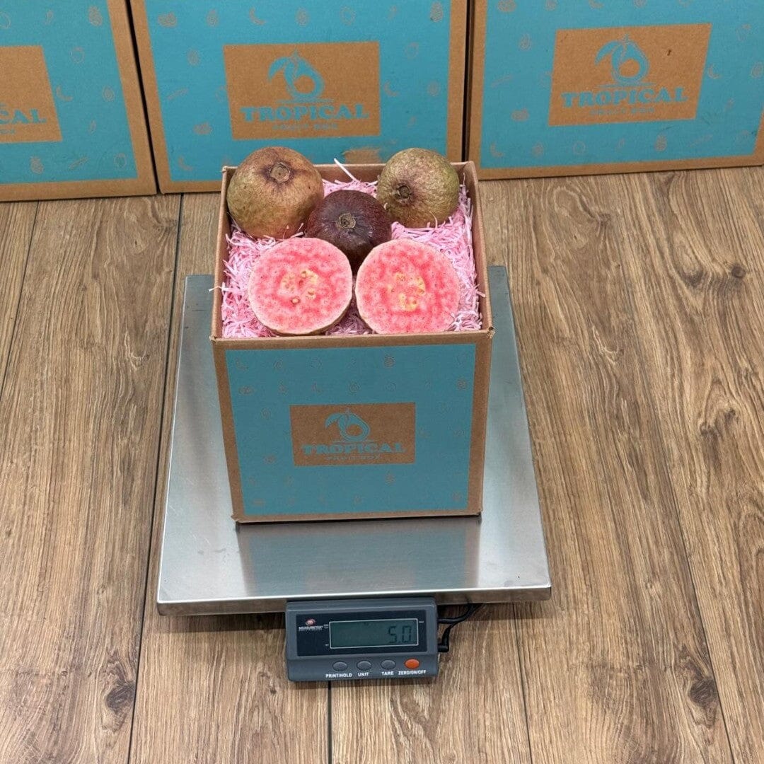 Tropical Fruit Box purple guava fruit, whole and halved, showing vibrant pink flesh and seeds. Pink shredded paper fills the box.